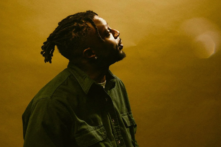 Profile portrait of rapper Isaiah Rashad in a green jacket looking upward against a warm yellow backdrop.